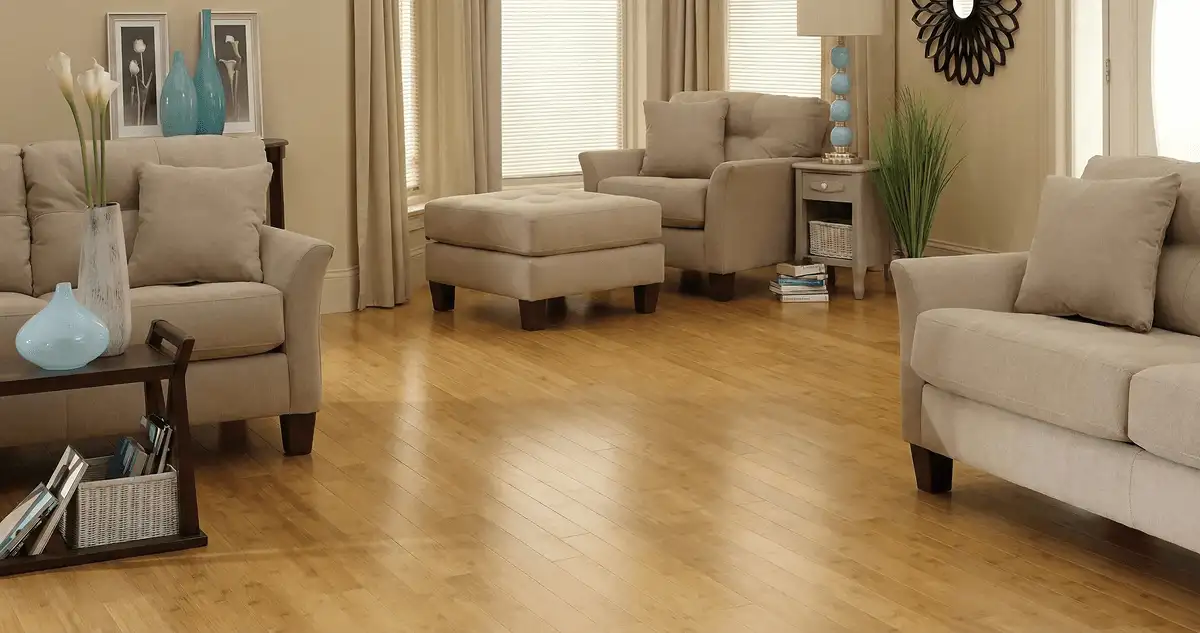 A living room with professionally installed bamboo flooring, beige sofas, a matching ottoman, a side table with a blue lamp, vases, framed pictures, and large windows with blinds and curtains in Nashua, NH