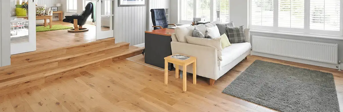 A modern living room with professionally installed laminate flooring, a beige sofa, a small side table, a gray rug, and large windows with white shutters in Nashua, NH
